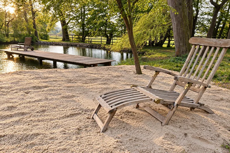 Ein Liegestuhl im Sand am Badeteich, dahinter der Holzsteg und der Park