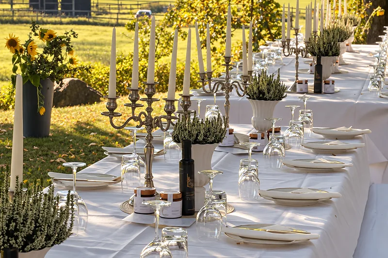 Eine lange weiße Tafel unter alten Bäumen, gedeckt mit Silberleuchtern, Kerzen und Gläsern